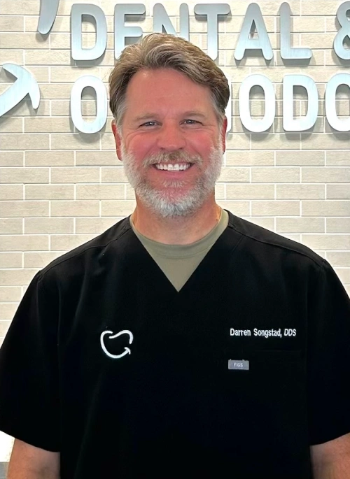Portrait of Dr. Darren Songstad, featuring a slightly wider view showing the tooth logo and "Darren Songstad, DDS" embroidered on his black scrubs. He stands smiling in front of a light gray brick office backdrop.