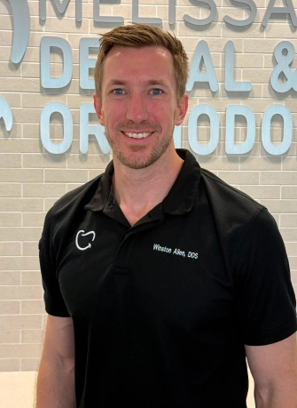 Portrait of Dr. Winston Allen, a man with short brown hair, short stubble facial hair, and blue eyes. He is wearing a black polo shirt embroidered with "Winston Allen, DDS" and a tooth logo. He is smiling in front of a light gray brick wall with metallic signage reading "...DENTAL & ORTHODONTICS."