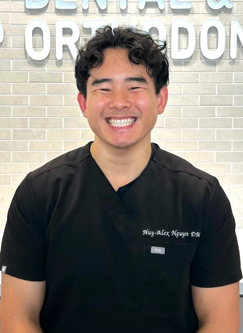 Portrait of Dr. Huy-Alex Nguyen, showing a wider view of his black dental scrubs embroidered with "Huy-Alex Nguyen, DMD." He is smiling widely against a light gray brick office wall.